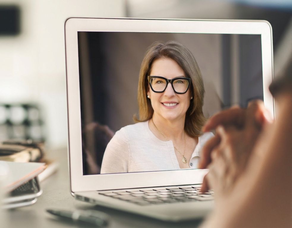 A laptop showing Dr. Jen Schafer on a video call, viewed over the shoulder of the person speaking with her.